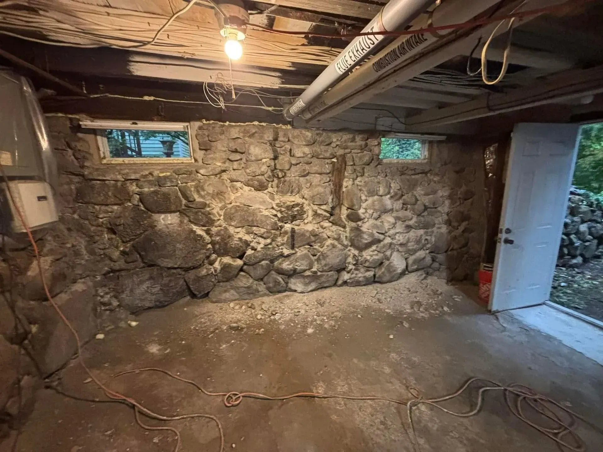Bare basement with stone wall, low windows, exposed ceiling, and concrete floor cluttered with cords