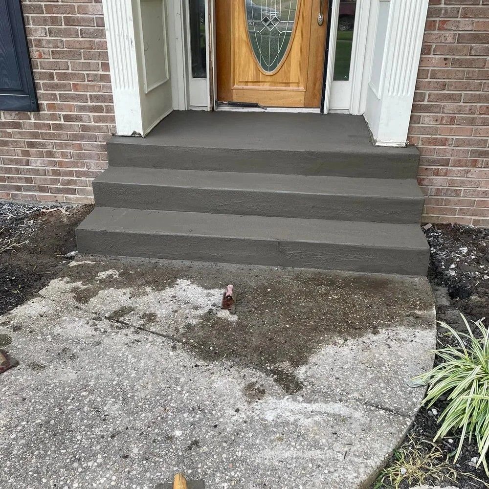 Front steps of a brick house with a wooden door, concrete walkway, and small landscaping bed.