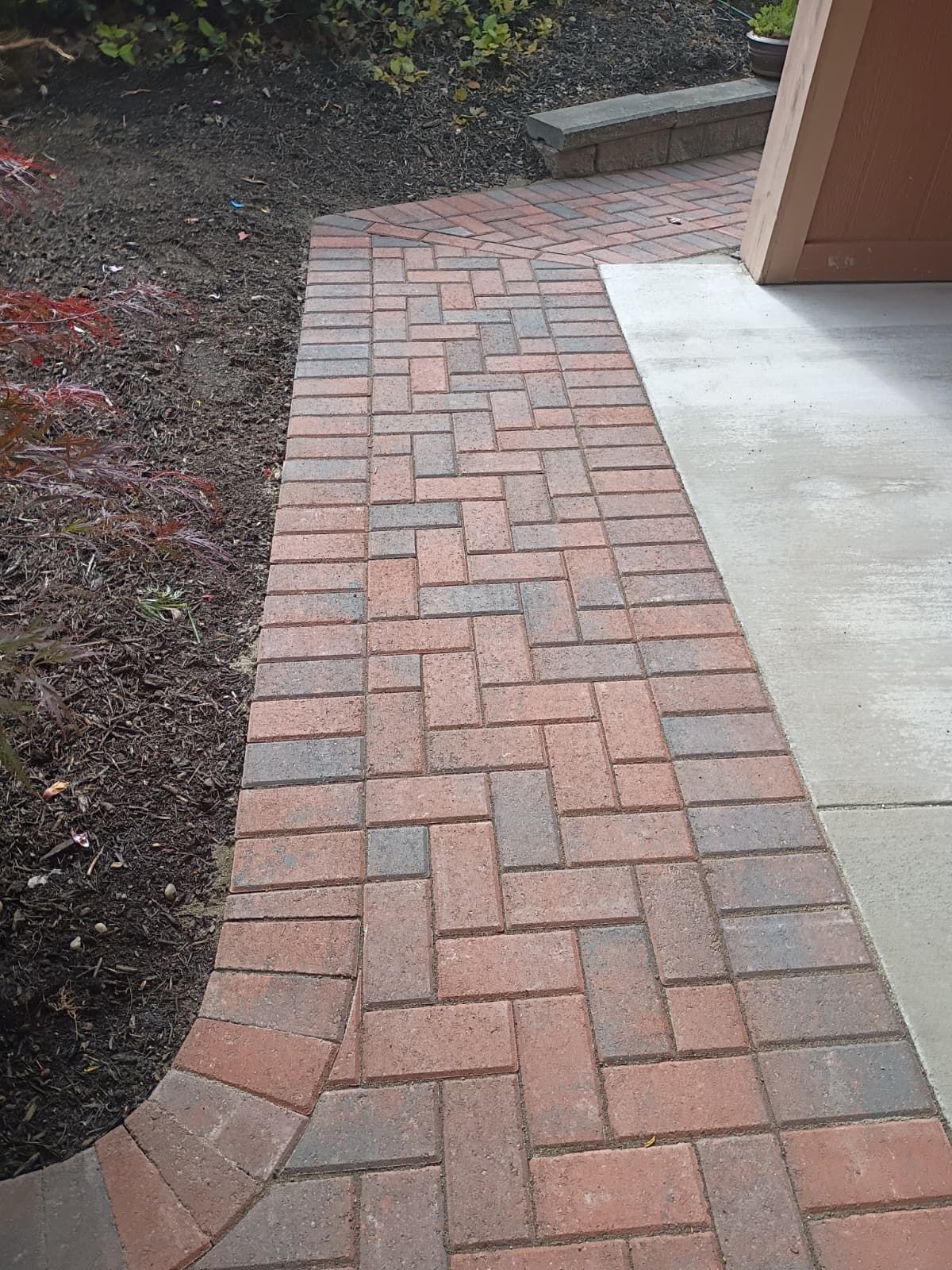 Brick paver walkway curving beside a concrete patio and garden bed