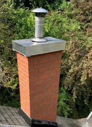 Red brick chimney with metal cap on a rooftop, set against green trees.