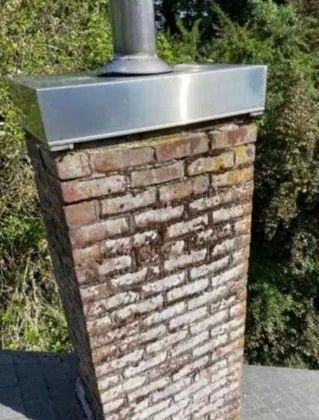 Brick chimney with a metal cap and flue against outdoor greenery.