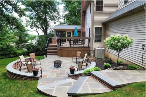 Outdoor stone patio with circular seating area, fire pit, and wooden deck beside a house and trees
