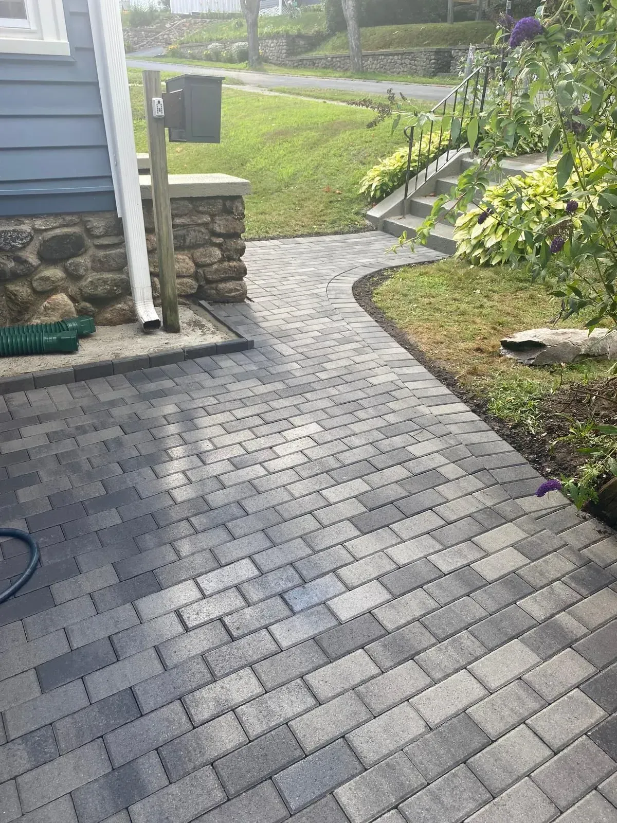 Brick walkway curving past a house and garden, with a mailbox and stone wall at the left.