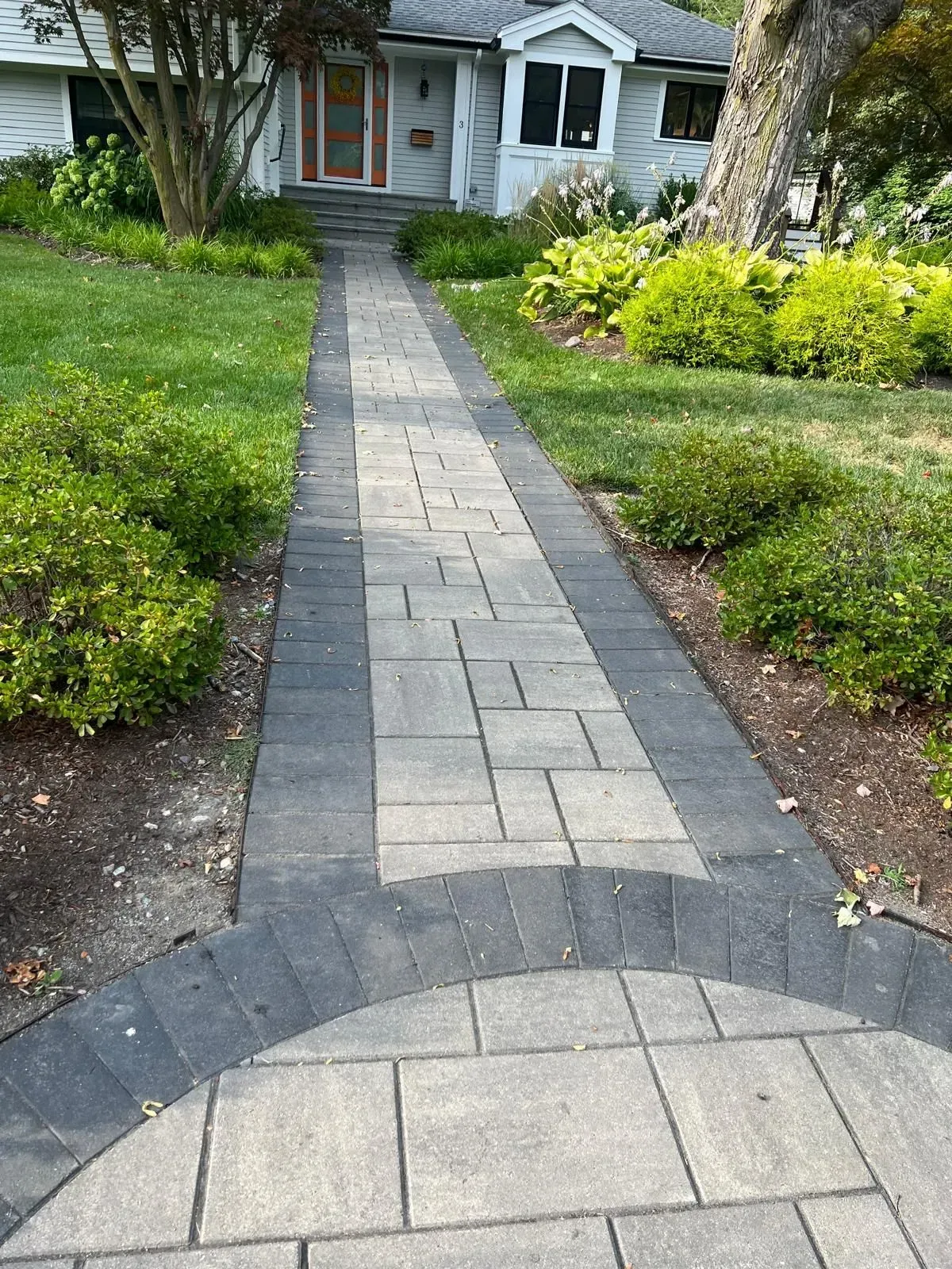 Paved walkway leading to a white house with a red door, bordered by green lawn and shrubs