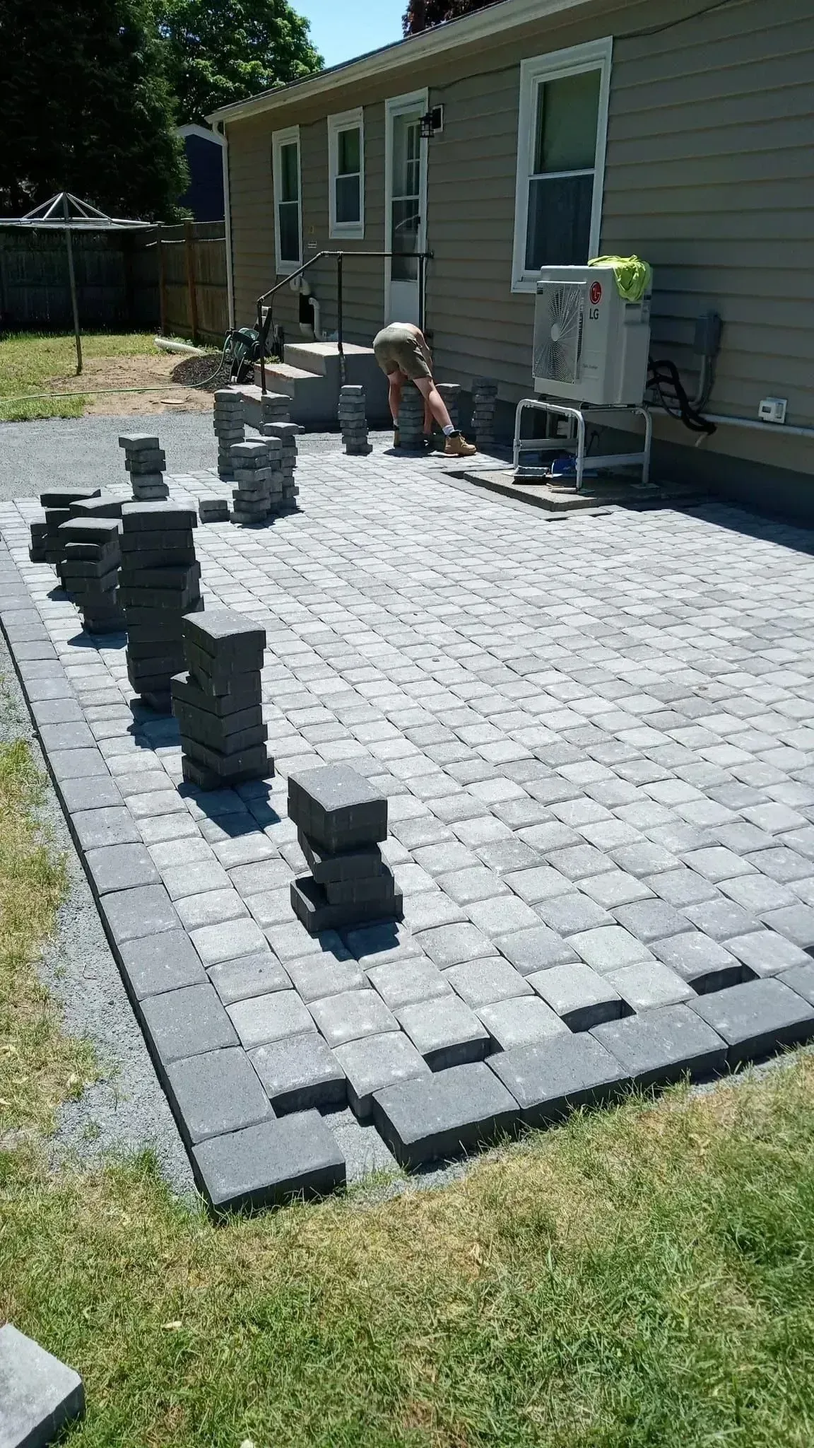 Backyard patio with gray pavers being laid beside a house, with stacked pavers and tools nearby