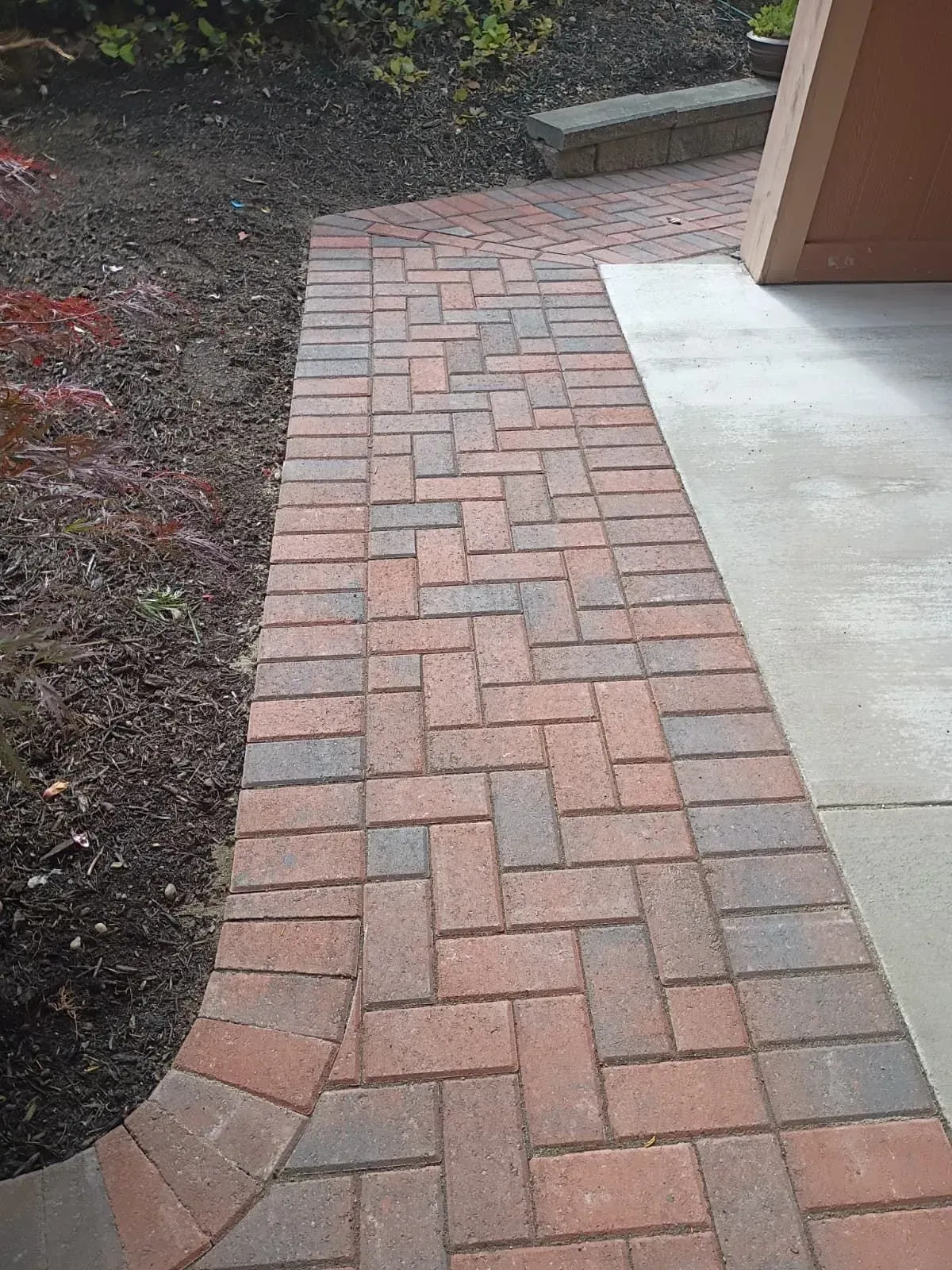 Brick walkway curves beside a porch, bordered by soil and a concrete slab.