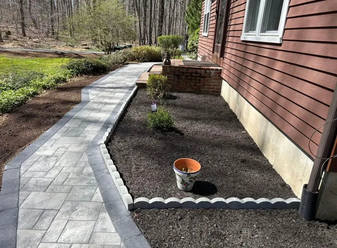 Curved garden walkway beside a house, with a mulched bed, scalloped edging, and a small orange bucket.
