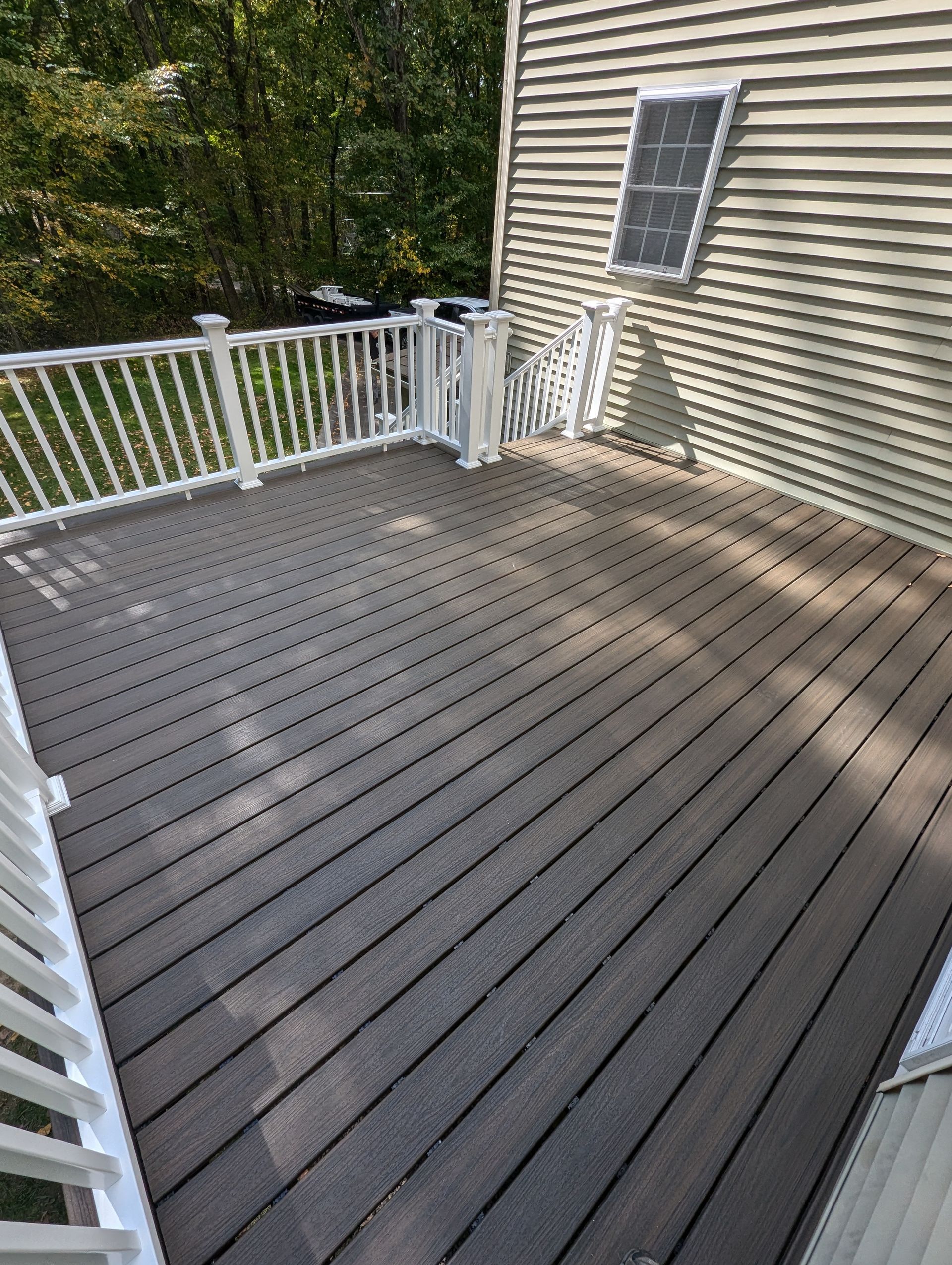 A gray composite deck with white railings, set against the side of a house with beige siding and a single window. 