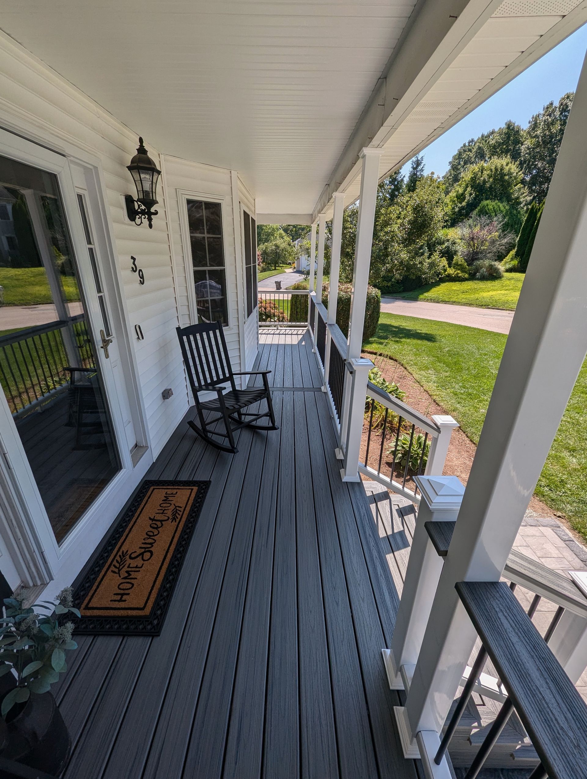 A front porch with grey decking, a black rocking chair, and a welcome mat outside a white house with a green lawn. 