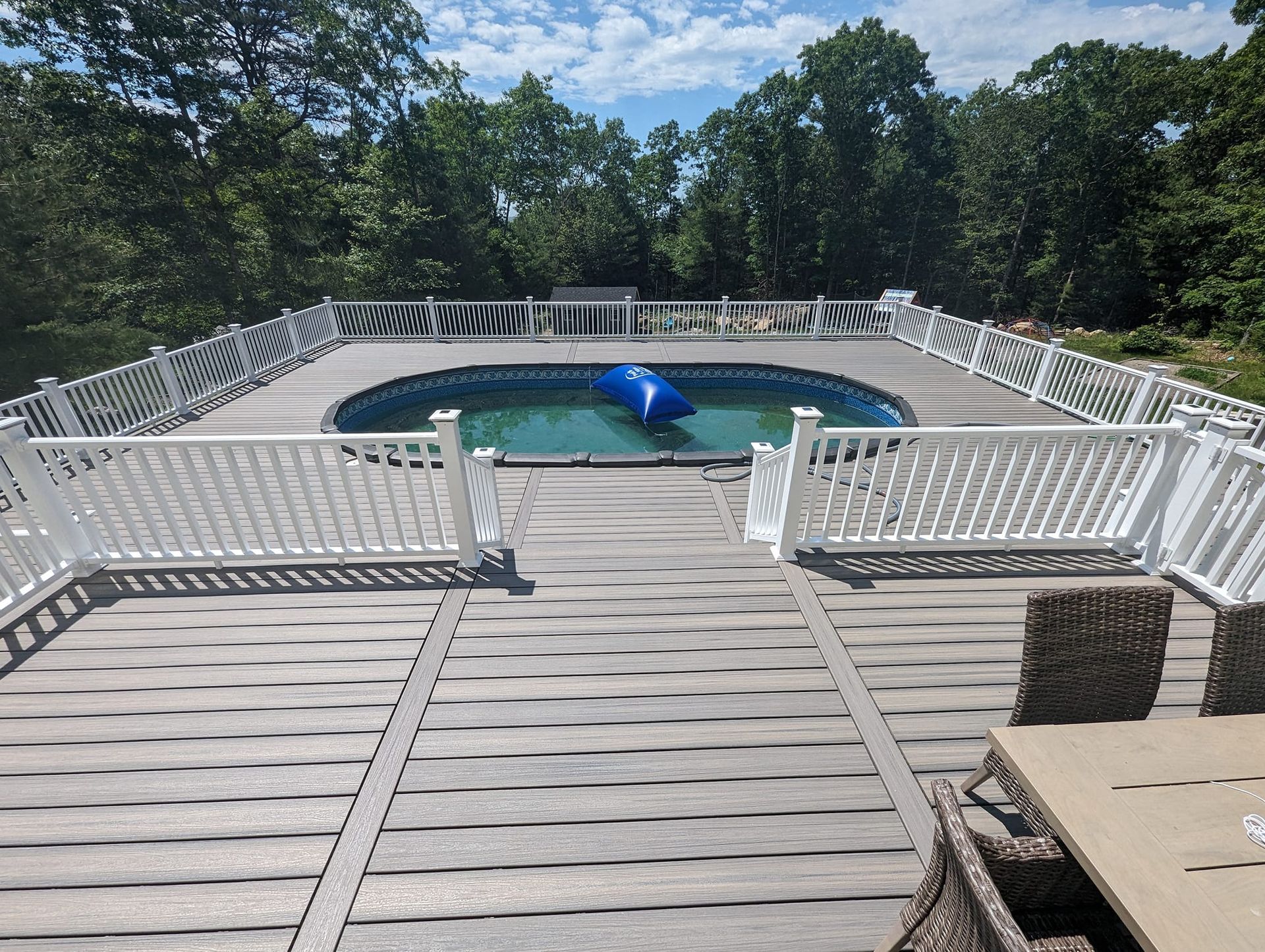 An above-ground pool surrounded by a light-gray composite deck and white railings, viewed from a wooden table.