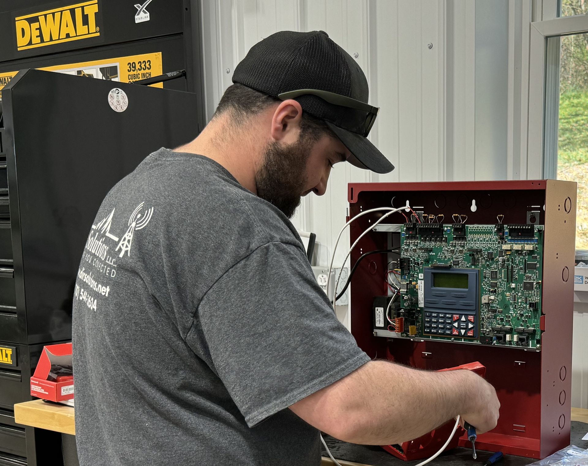 Man in gray shirt installing circuit board in a red metal box, wearing a hat, inside a workshop.
