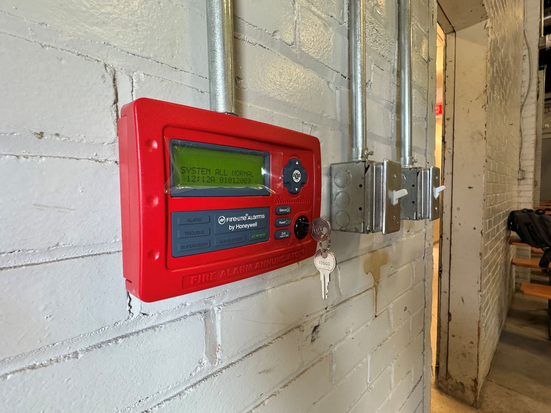 Red fire alarm panel on a white brick wall with a key, metal conduit, and electrical boxes.