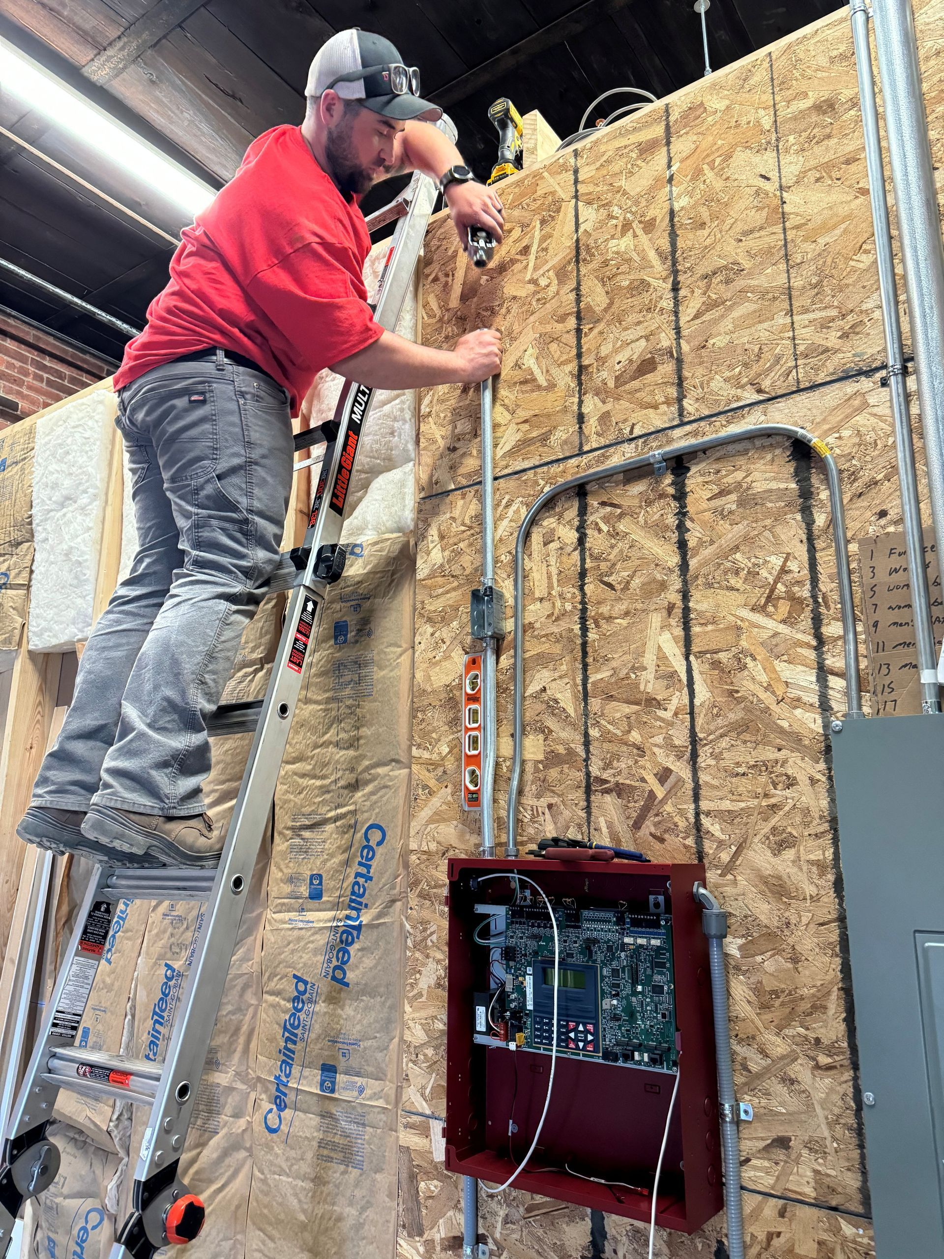 Man in red shirt on ladder installing electrical conduit on a plywood wall near a red control box.