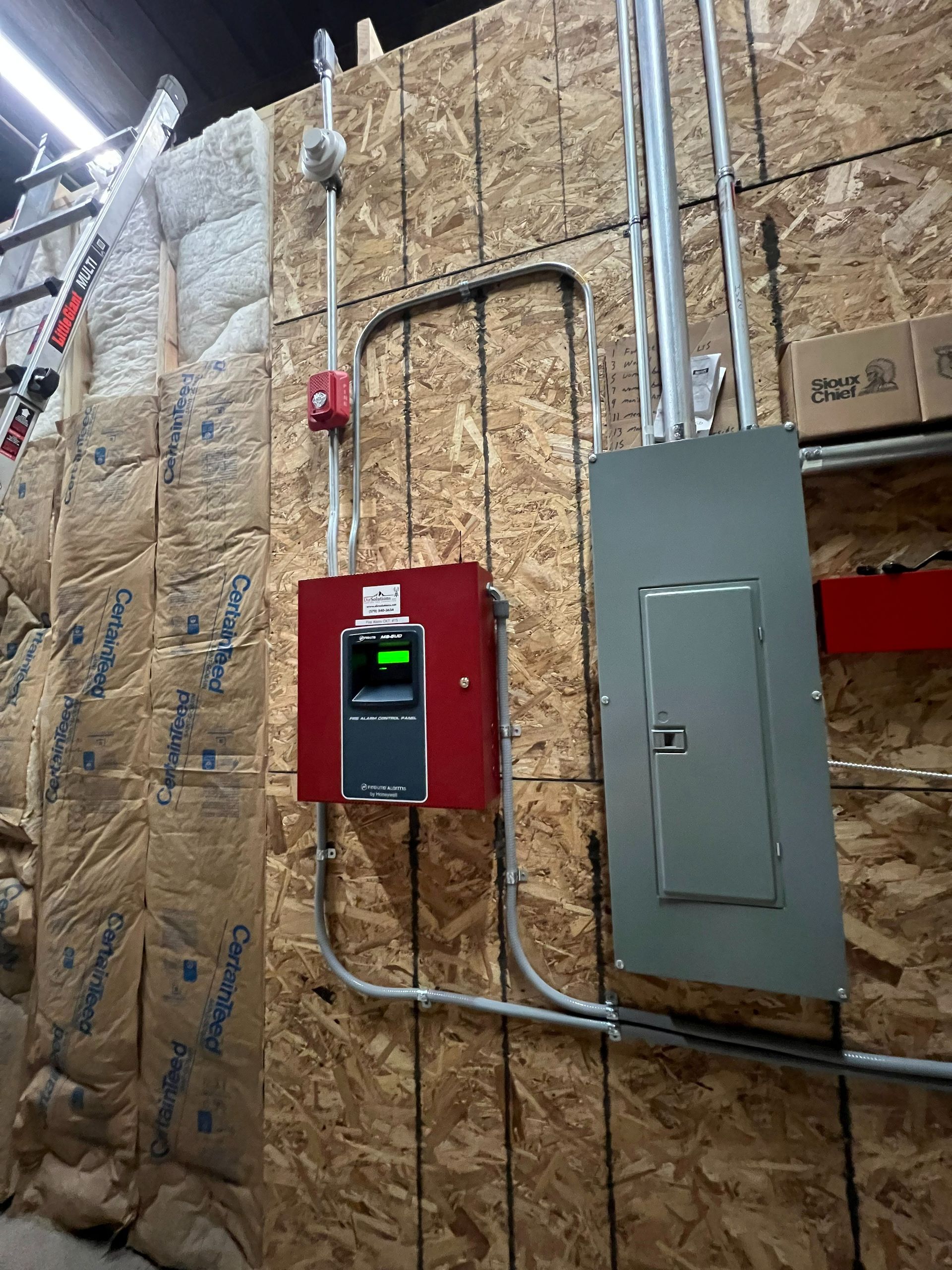 Fire alarm system components mounted on a wood-paneled wall, including a red panel, pull station, and electrical panel.