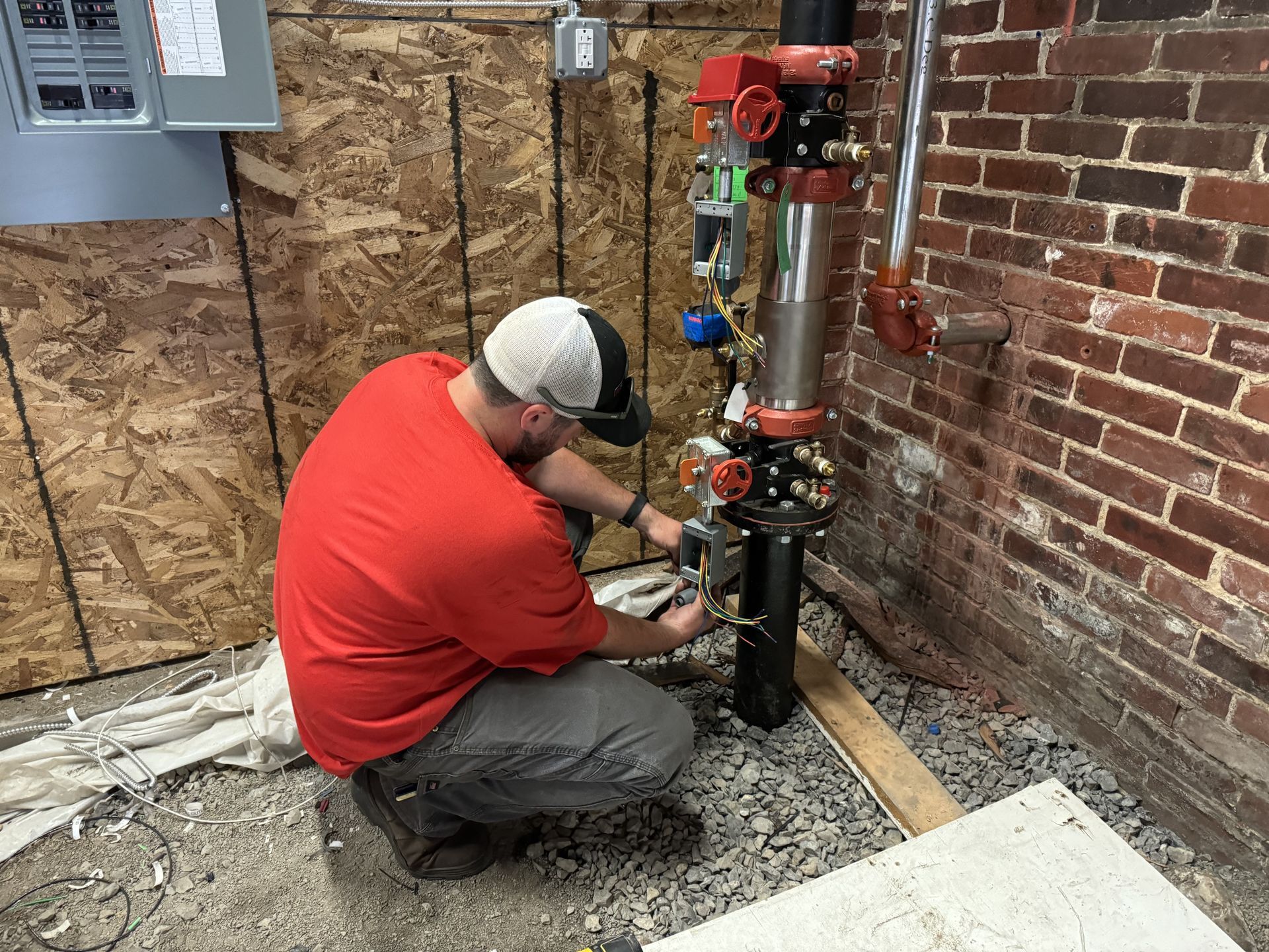 Man in red shirt working on plumbing near brick wall.