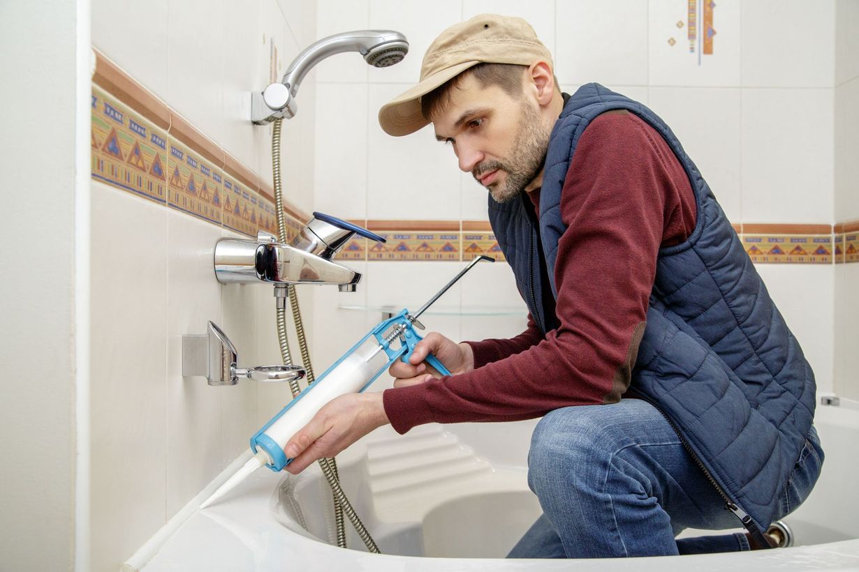 Person kneeling by a bathtub, using a caulking gun to seal the shower area.