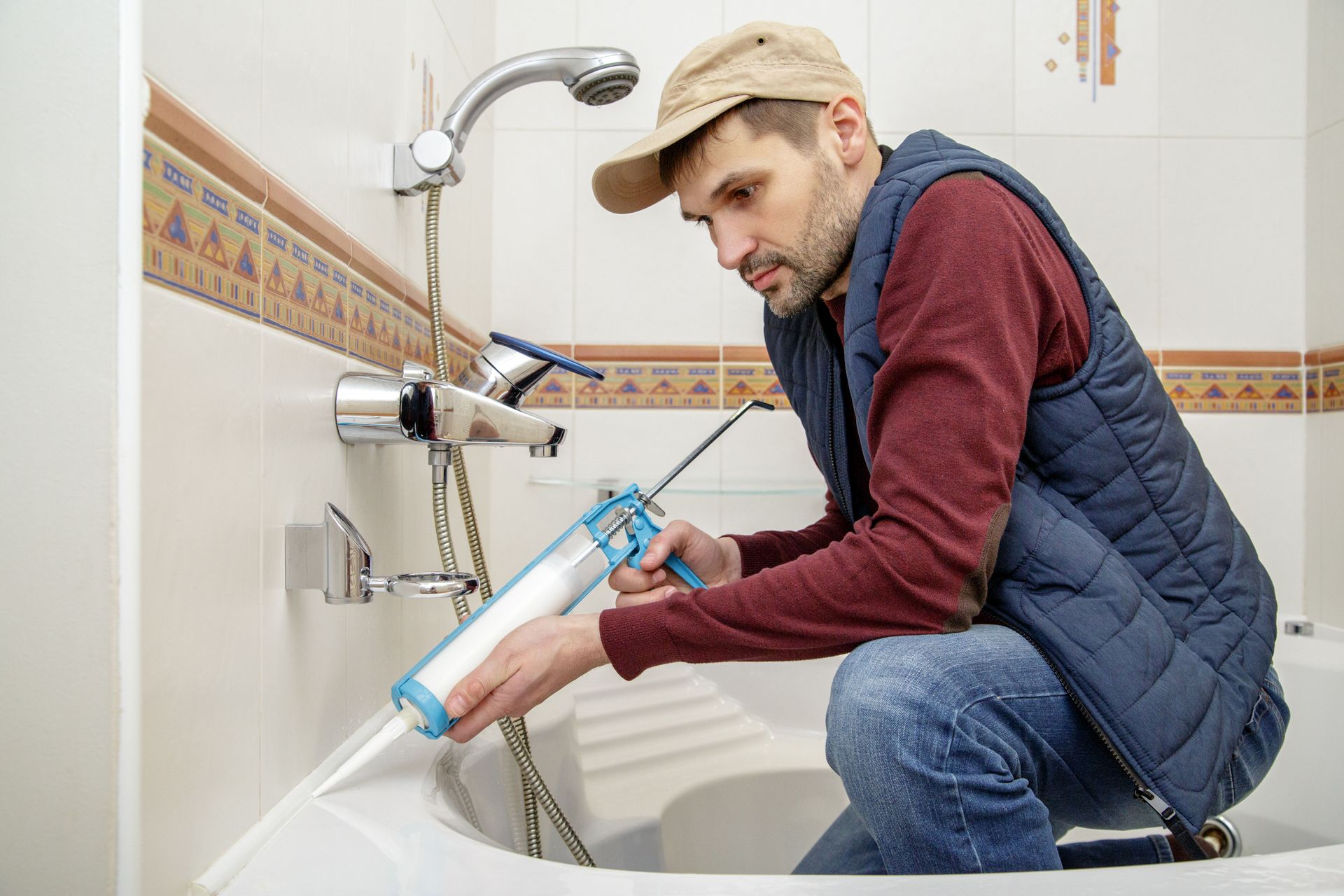 Person kneeling by a bathtub, using a caulking gun to seal the shower area.