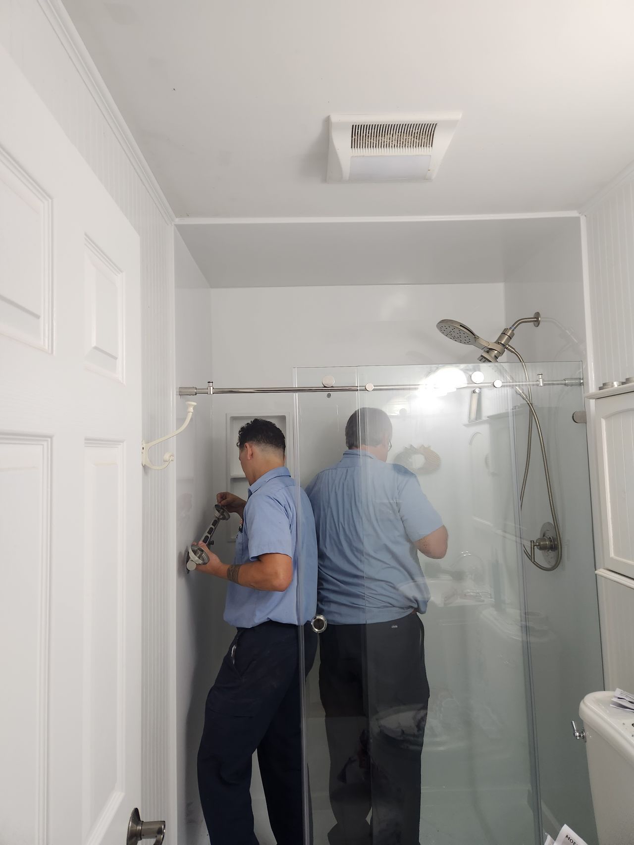 Technician working in a bathroom beside a glass shower stall and sink, seen in a mirror.