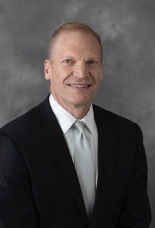 Studio portrait of a man in a black suit, white shirt, and light tie, smiling against a gray background