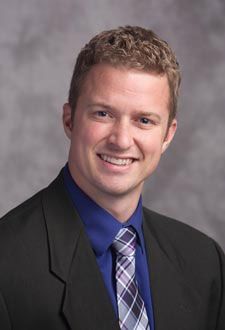 Smiling man in a dark suit and blue shirt with a plaid tie against a gray studio backdrop