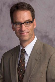 Portrait of a man in glasses wearing a tan suit and patterned tie against a gray studio backdrop