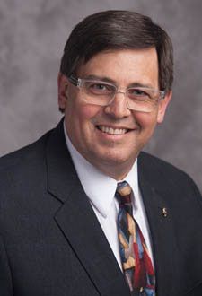 Smiling man in a dark suit, glasses, and patterned tie against a gray studio backdrop