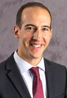 Smiling man in a dark suit with a white shirt and burgundy tie against a gray background