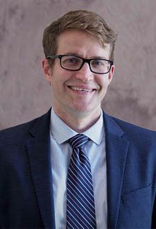 Smiling man in a blue suit, striped tie, and glasses against a neutral backdrop