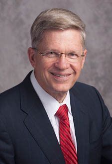 Portrait of a smiling man in a dark suit, white shirt, and red tie against a gray backdrop