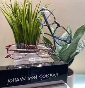 Three colorful eyeglass frames displayed on a stand with green plants in the background.