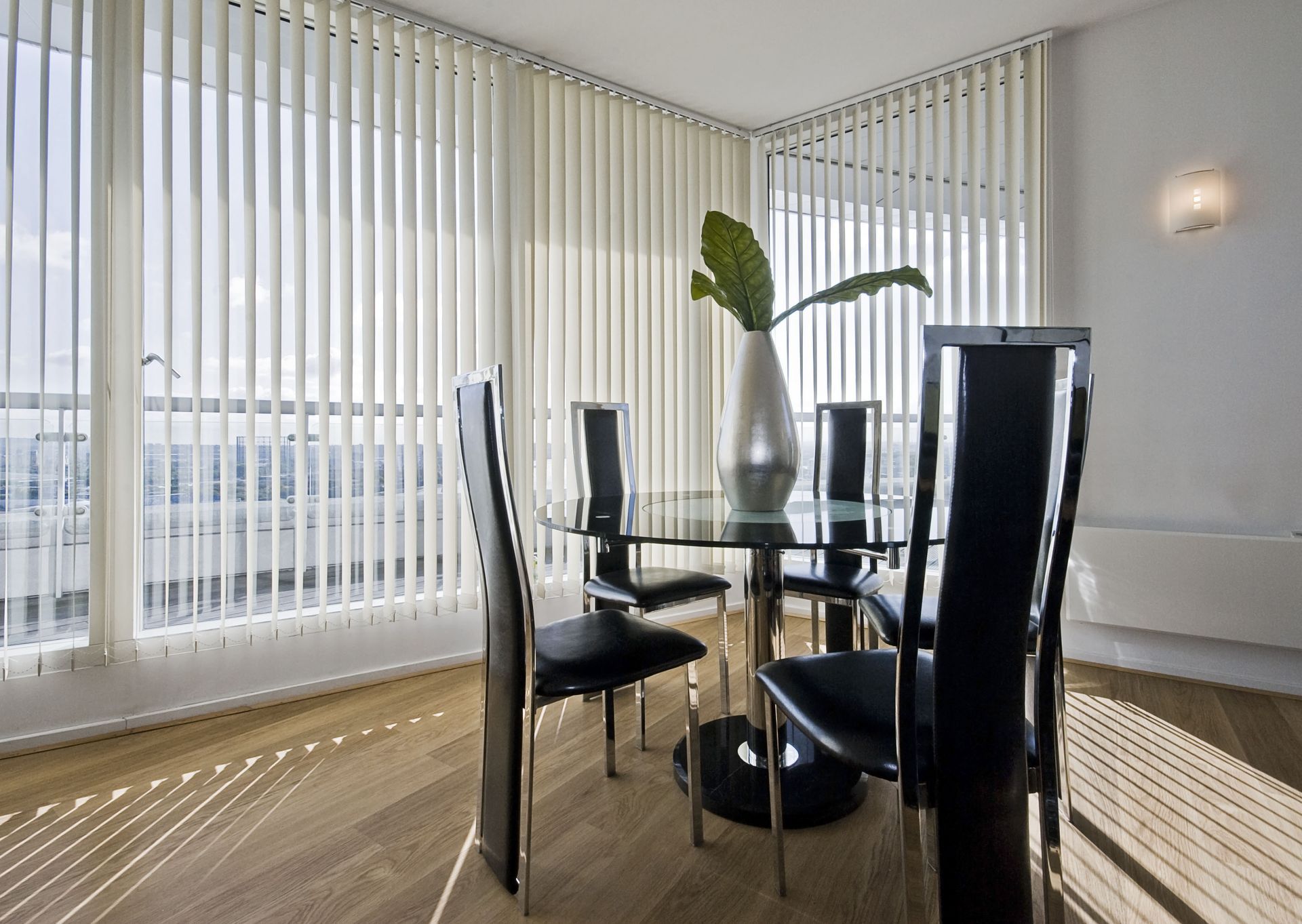 Dining room with glass table, black chairs, and vertical blinds. A vase with a plant sits on the table.