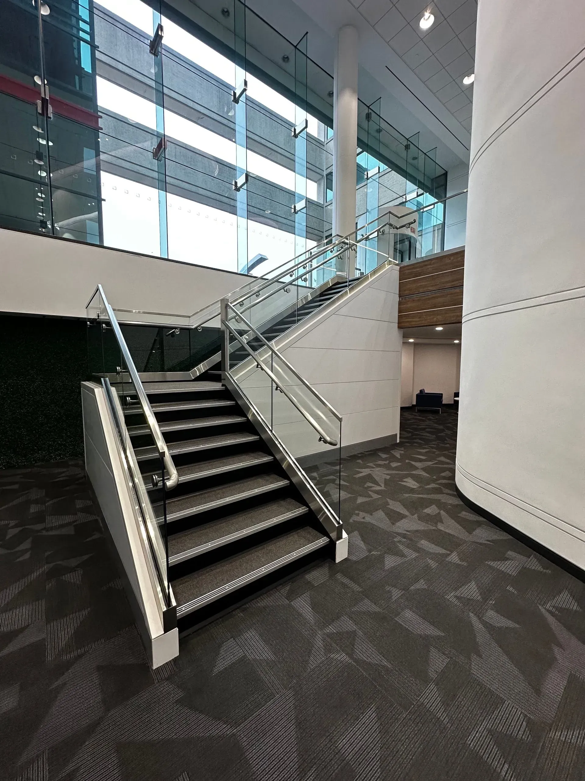 Staircase with glass railing, ascending into a modern building. Black and grey carpet. White walls.
