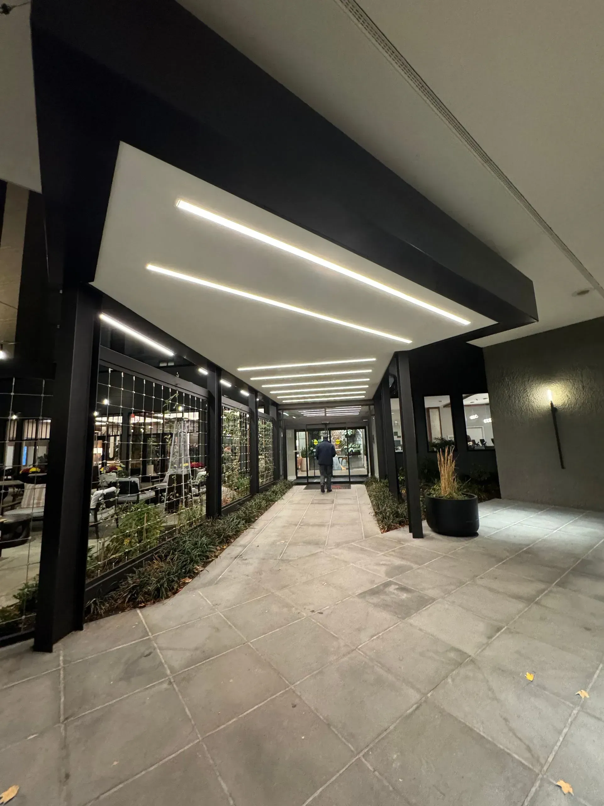 Covered walkway entrance with linear lights. A person walks toward the door, surrounded by dark beams and greenery.