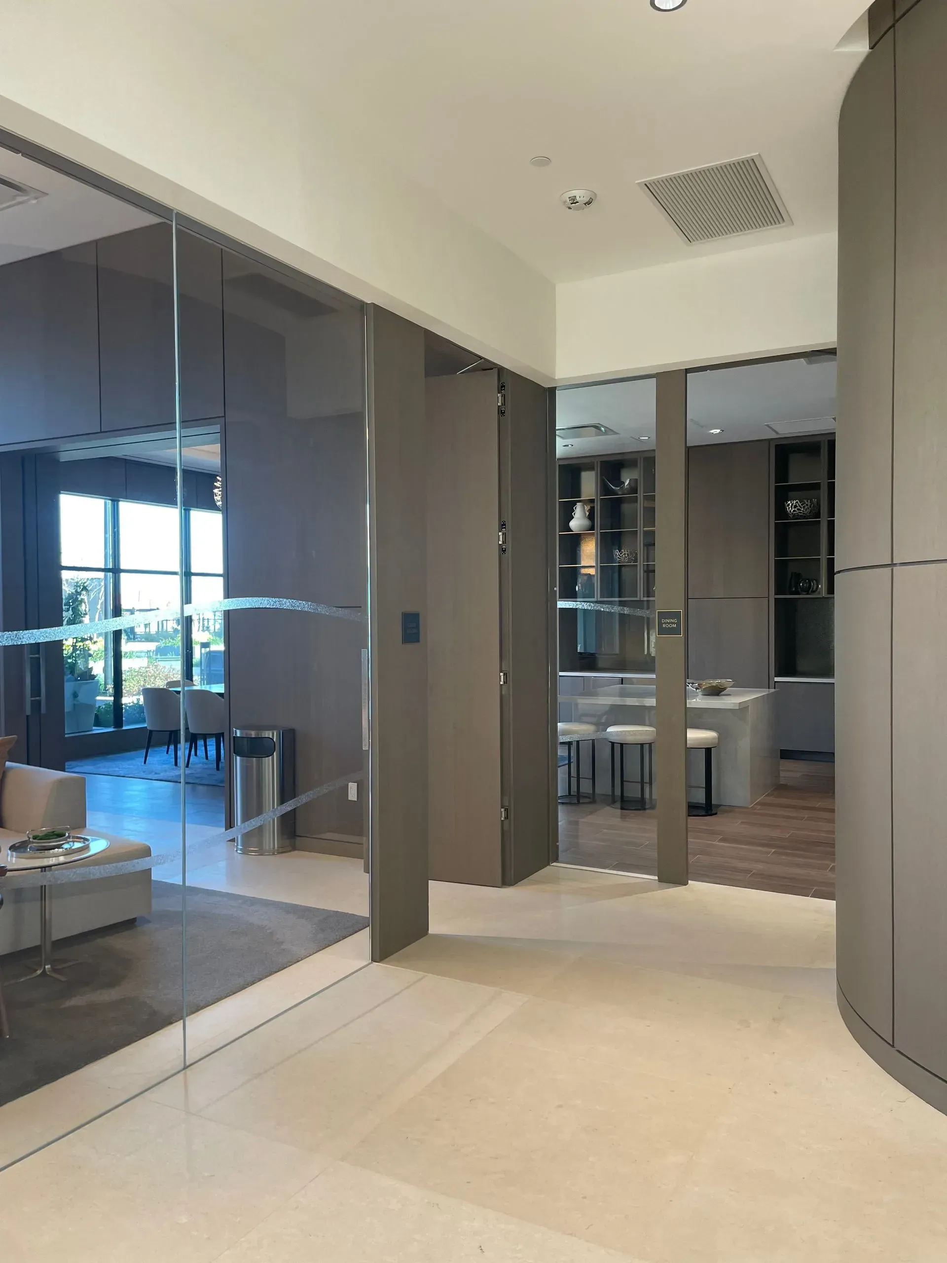 Hallway with glass walls and neutral tones leading to a living room and kitchen.