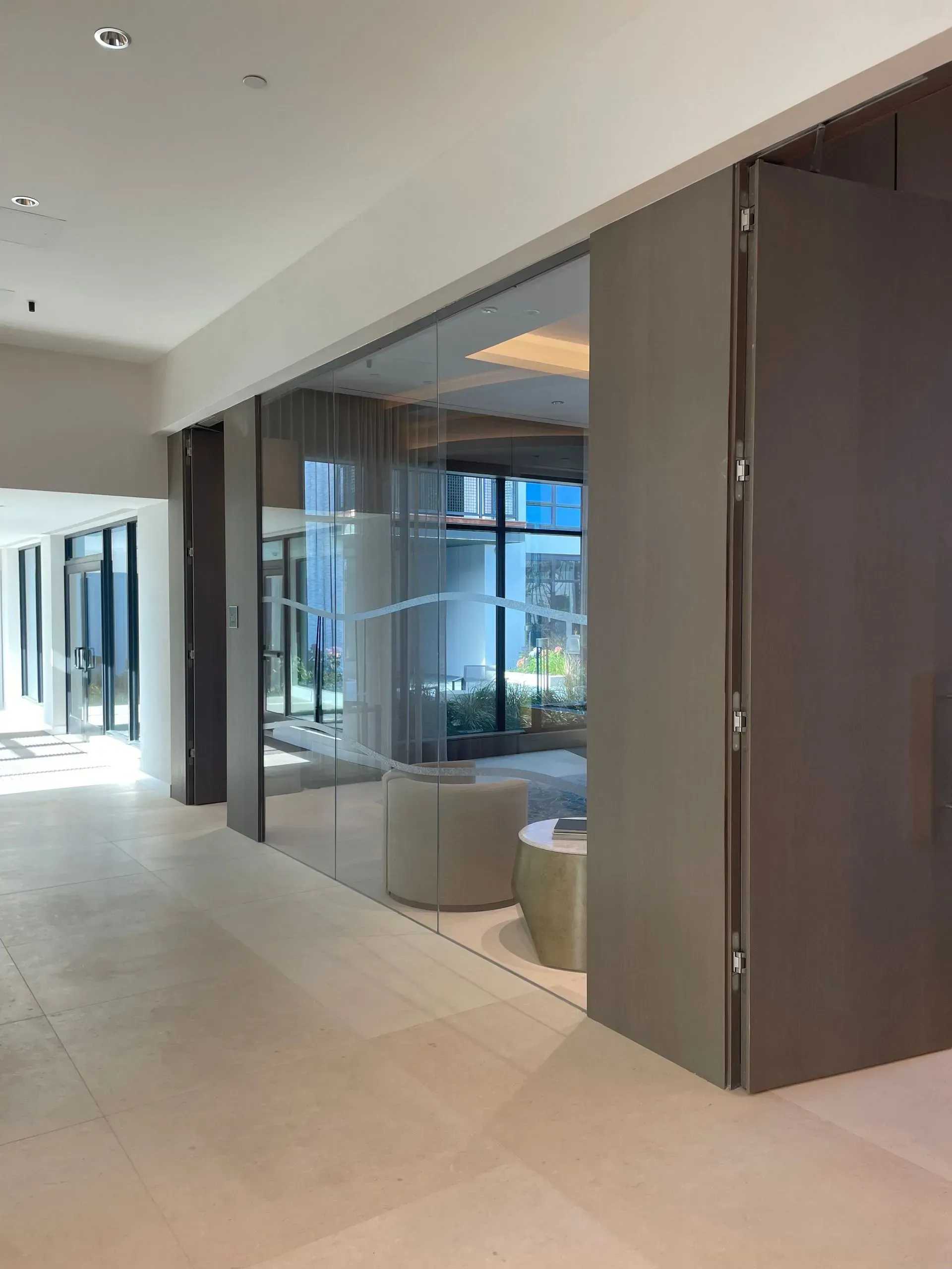Glass-walled office space with beige walls and flooring; large folding door, open to a waiting area with neutral furniture.