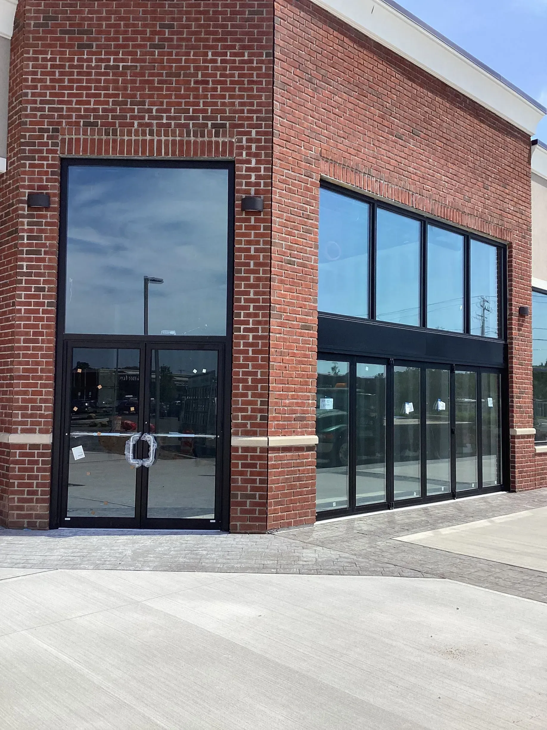Red brick building with large glass doors and windows, black frames. Concrete pavement in foreground.