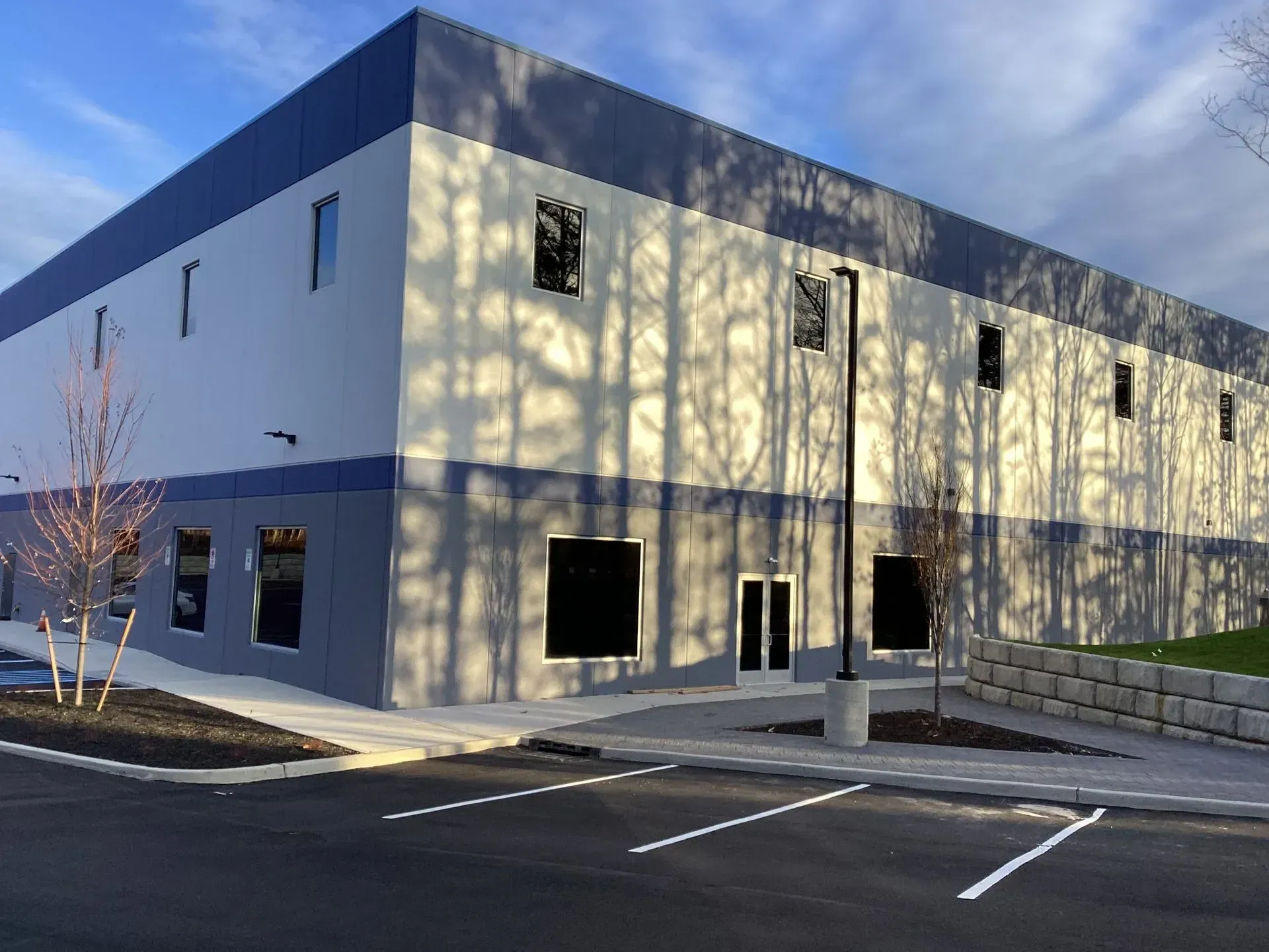 Industrial building with blue and gray exterior, trees casting shadows, and parking spaces.