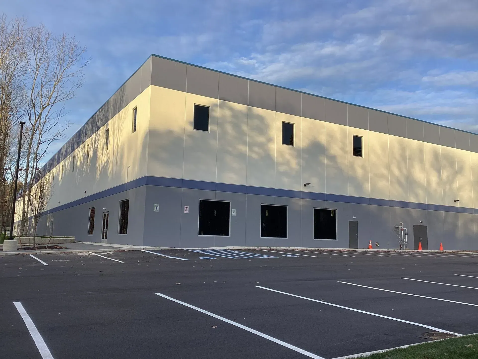 Exterior view of a large industrial building with a light beige and gray facade, and a paved parking lot.