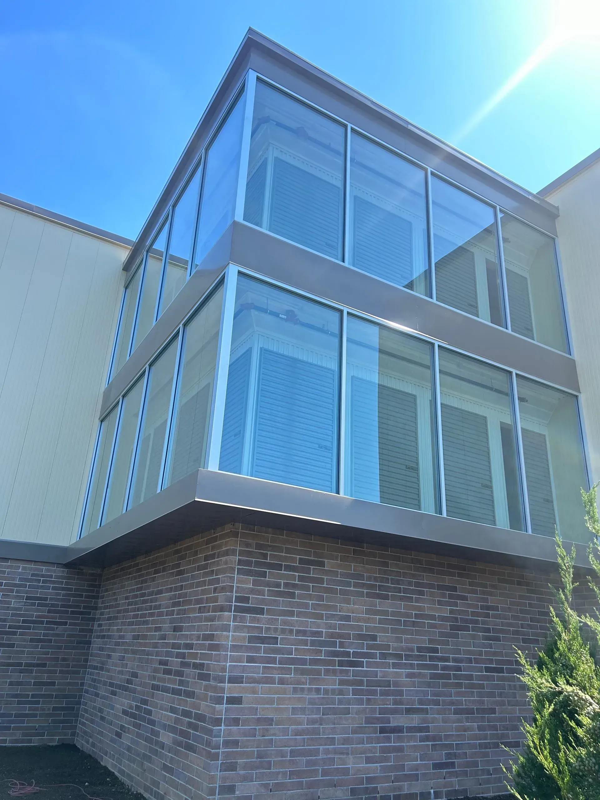 Two-story glass-walled addition over a brick base attached to a light beige building against a clear blue sky.