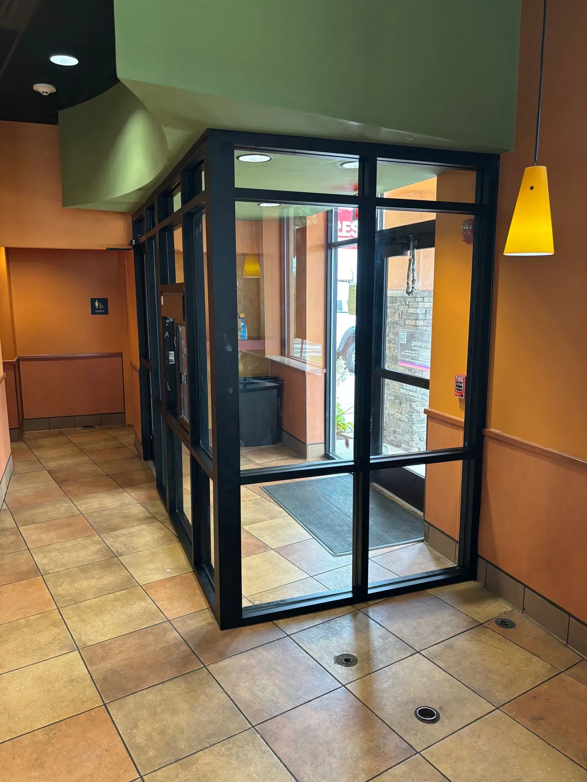 Restaurant entrance with glass doors framed in black. Orange walls, tiled floor, and a yellow light fixture are visible.