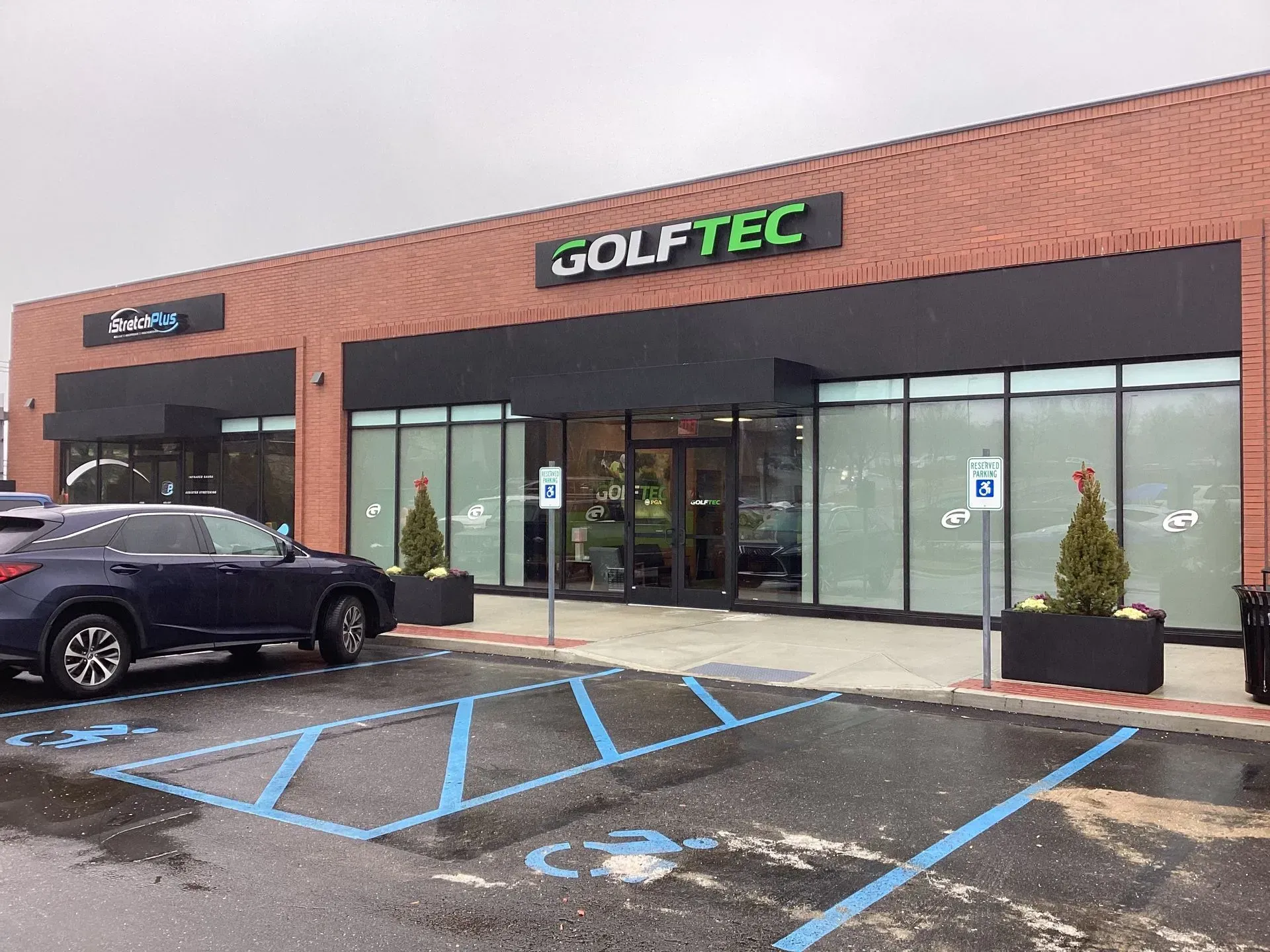 Exterior of a Golftec store with brick facade, large windows, and parking spots. A blue car is parked in the lot.