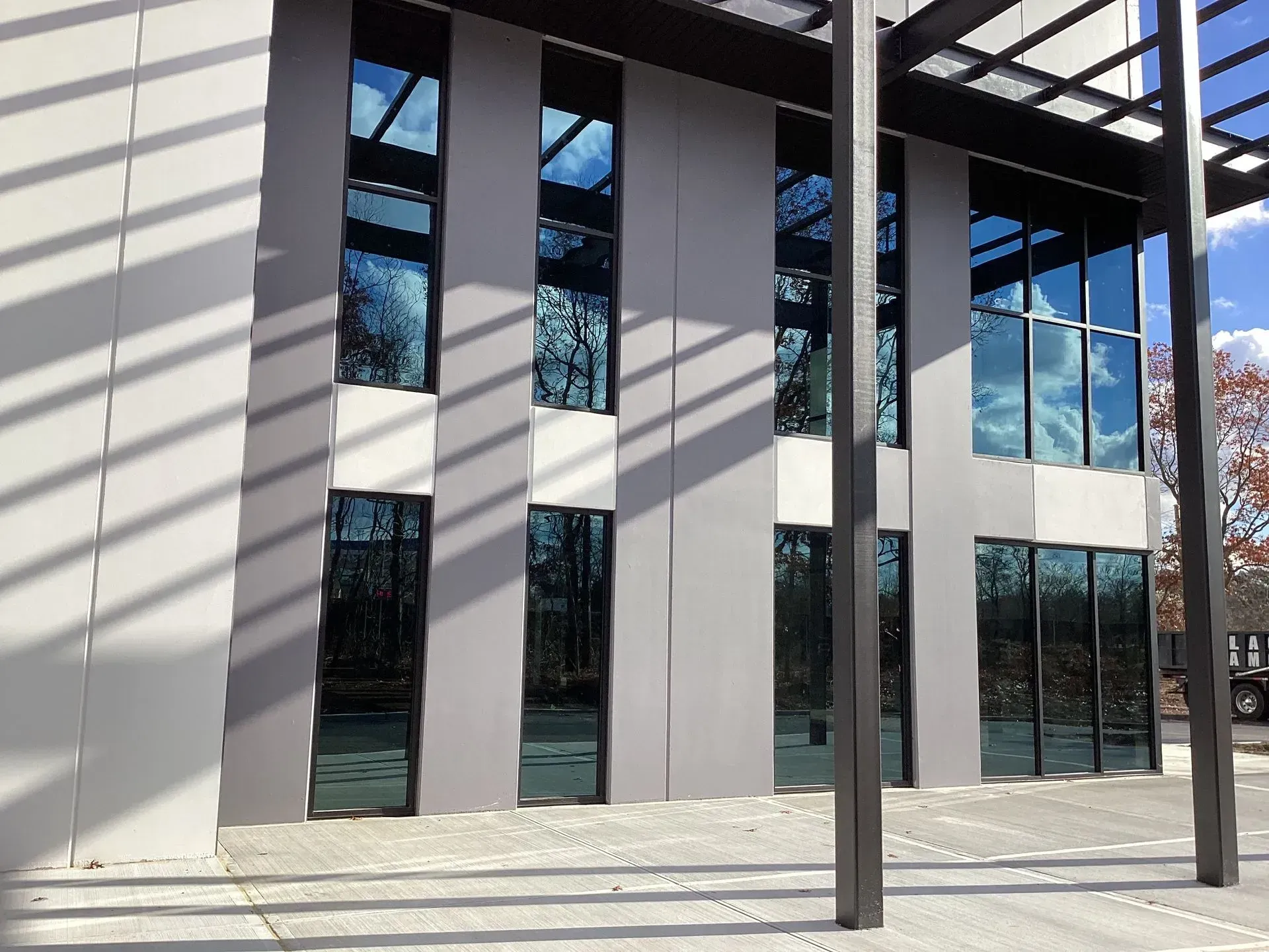 Modern building with tall windows reflecting a blue sky and trees. Gray exterior, metal supports.