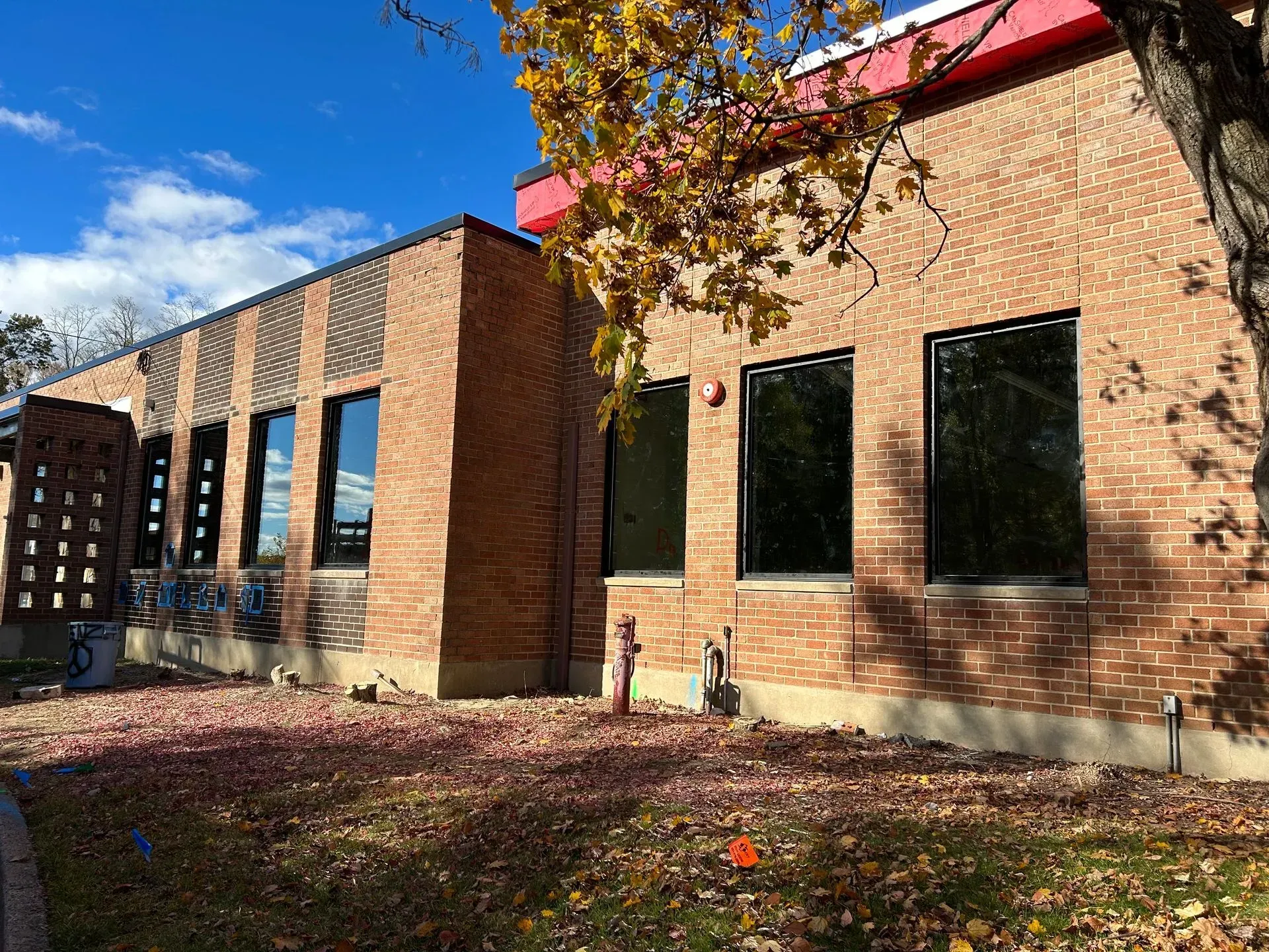 Brick building with black-framed windows and a red roof trim. Autumn leaves are on the ground.