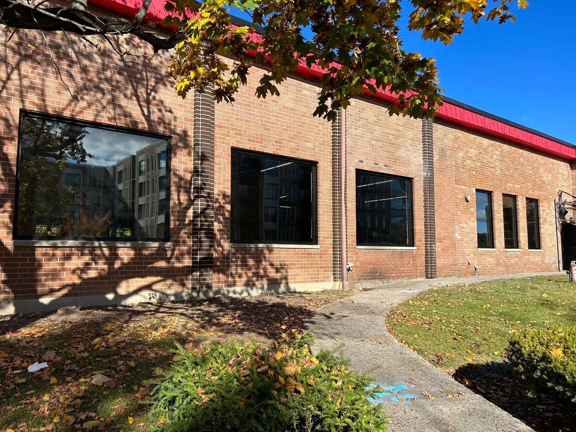 Brick building with large black-framed windows, red roof, and a curved pathway. Fall foliage and sunny day.