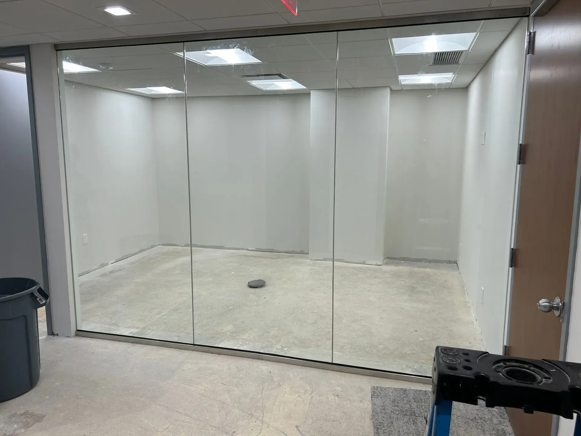 Glass-walled office with white walls and ceiling, empty. A trash can and tool are in the foreground.
