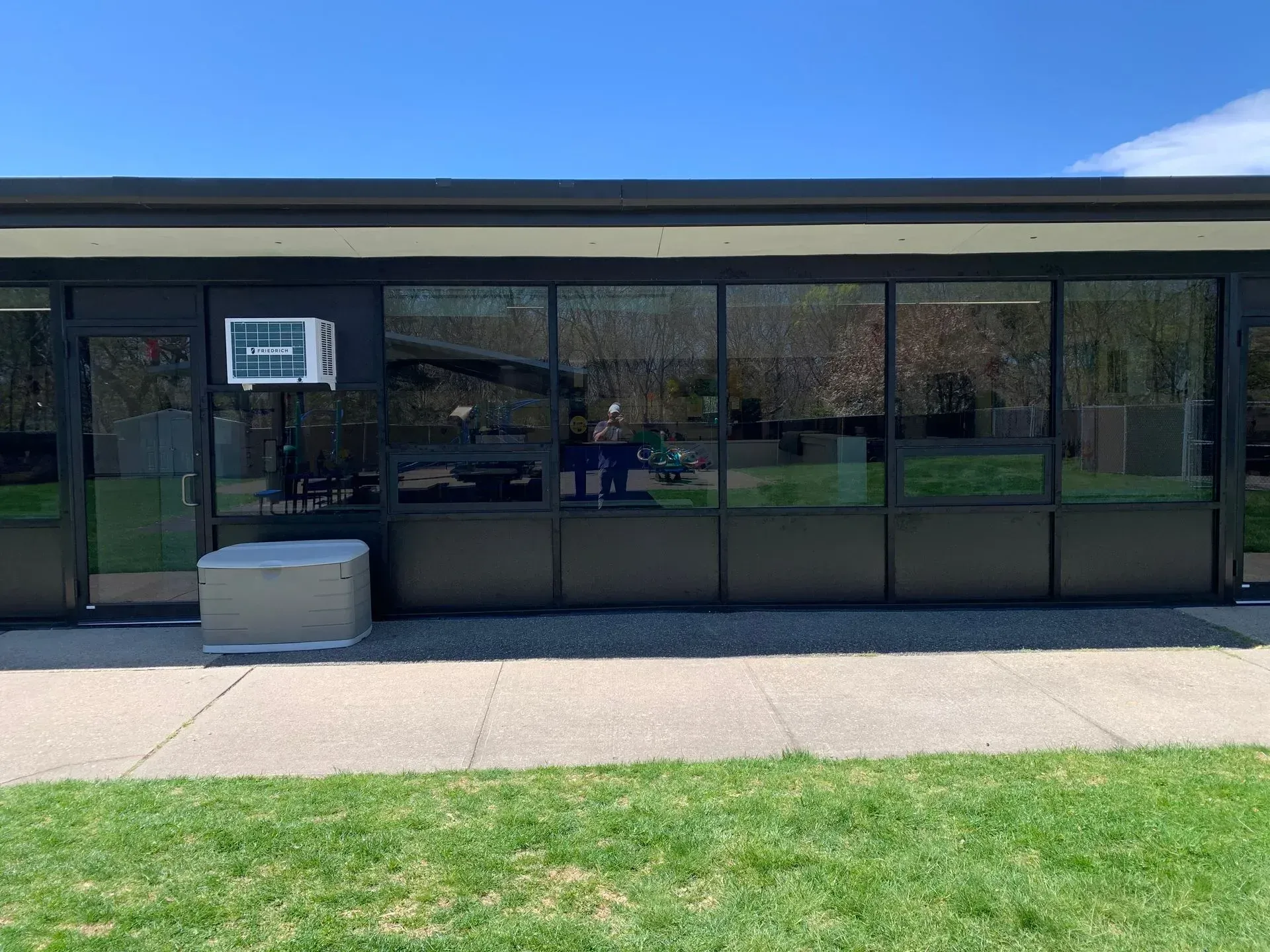 Building with black-framed windows reflecting trees, with an air conditioner and large box on the side, on a sunny day.
