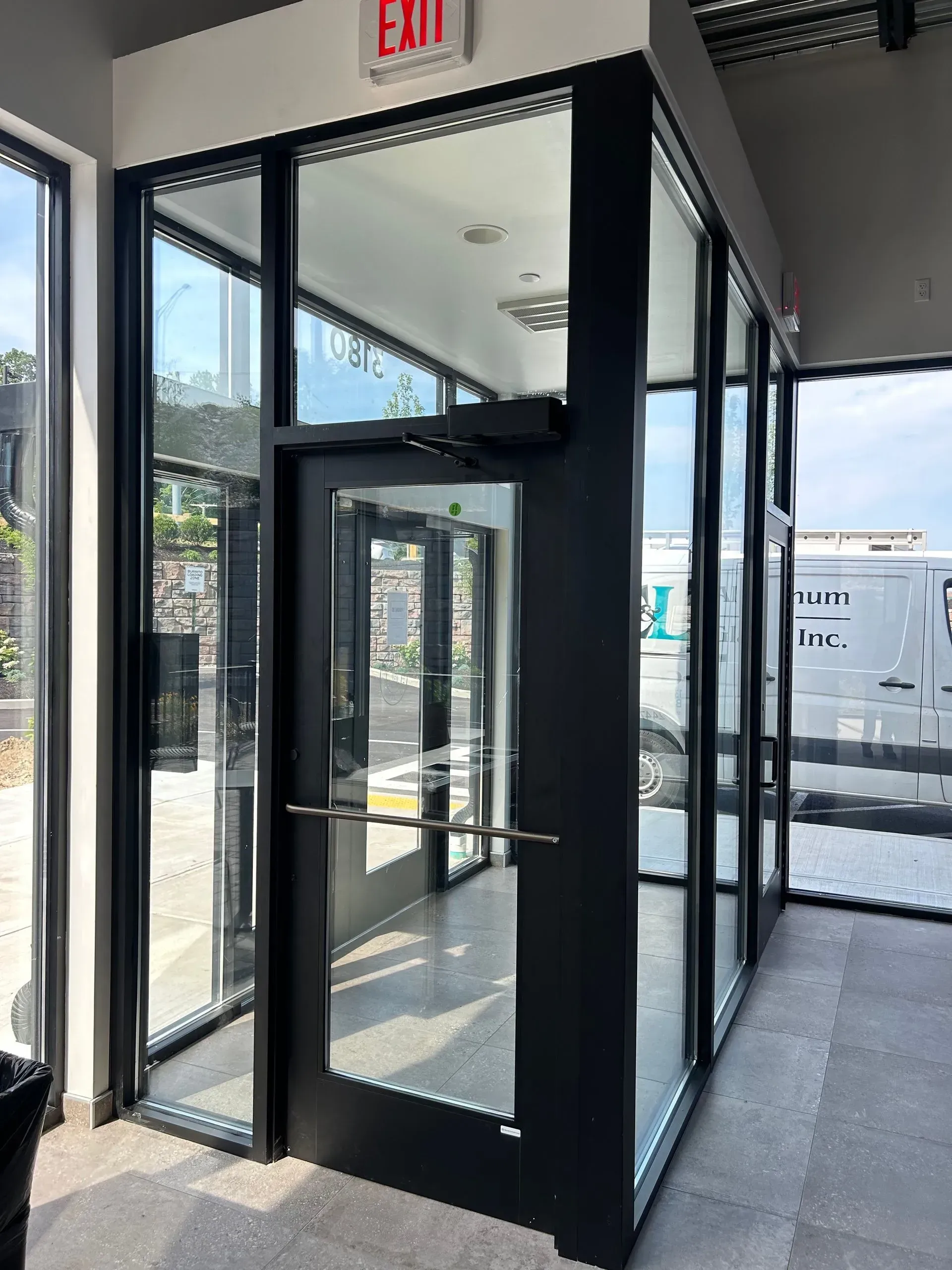 Glass-walled hallway with dark trim and an automatic door. Exit sign above. Grey floor, outdoor view.