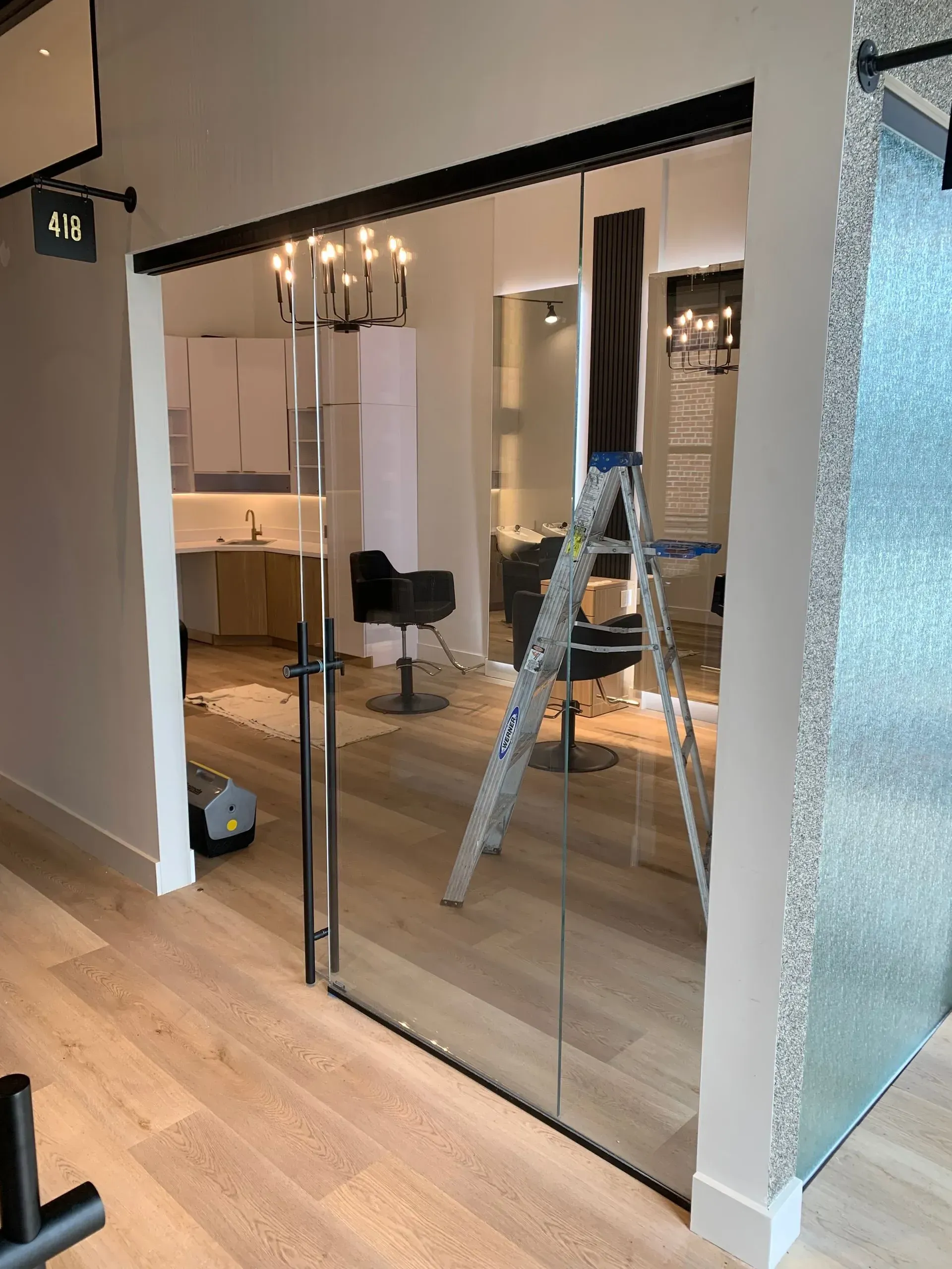 Glass door with black frame, opens into a salon with chairs, mirrors, and ladder on wood floor.