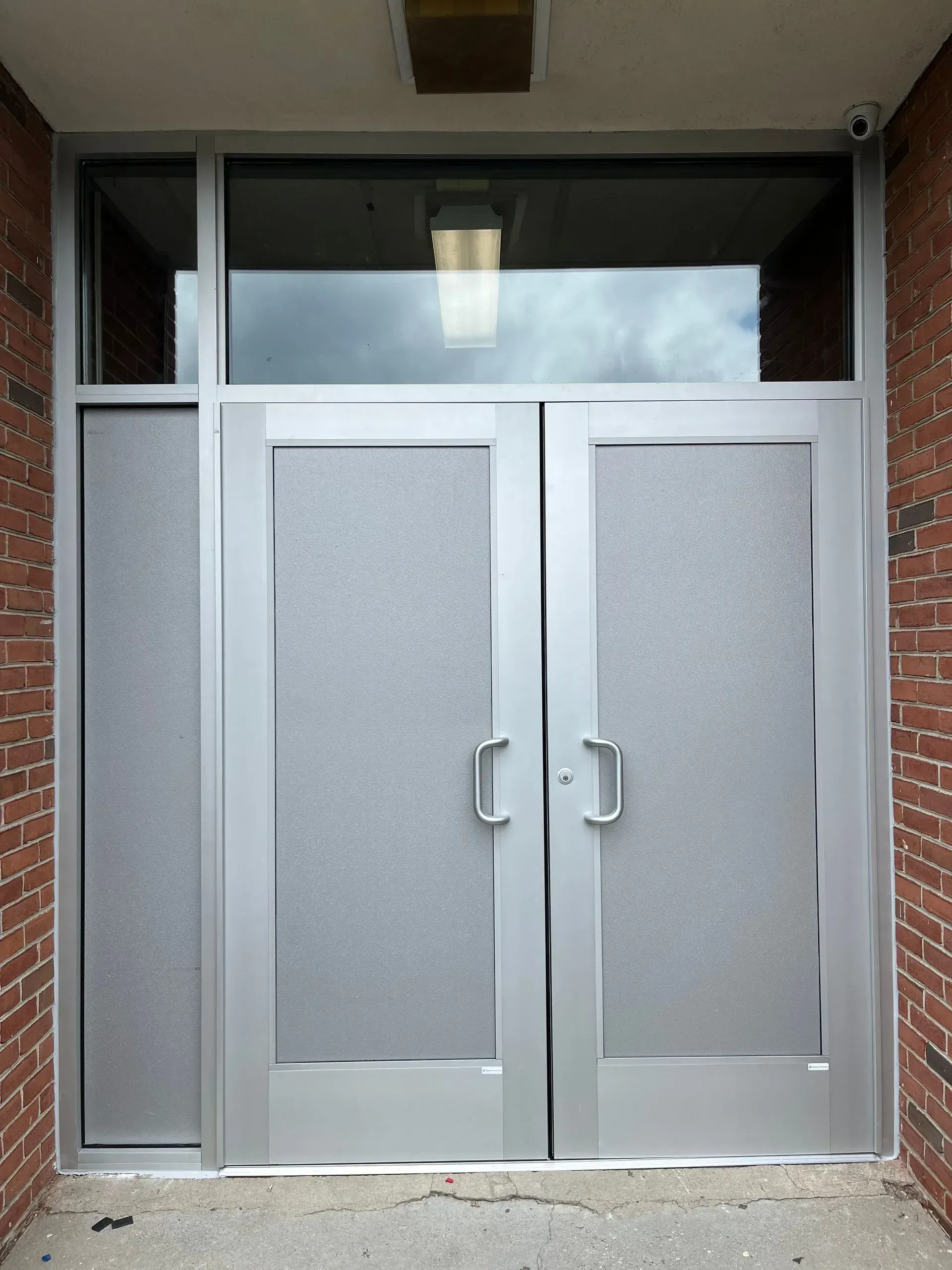 Double metal doors with frosted glass, side panel, and transom, framed in silver, set in brick.