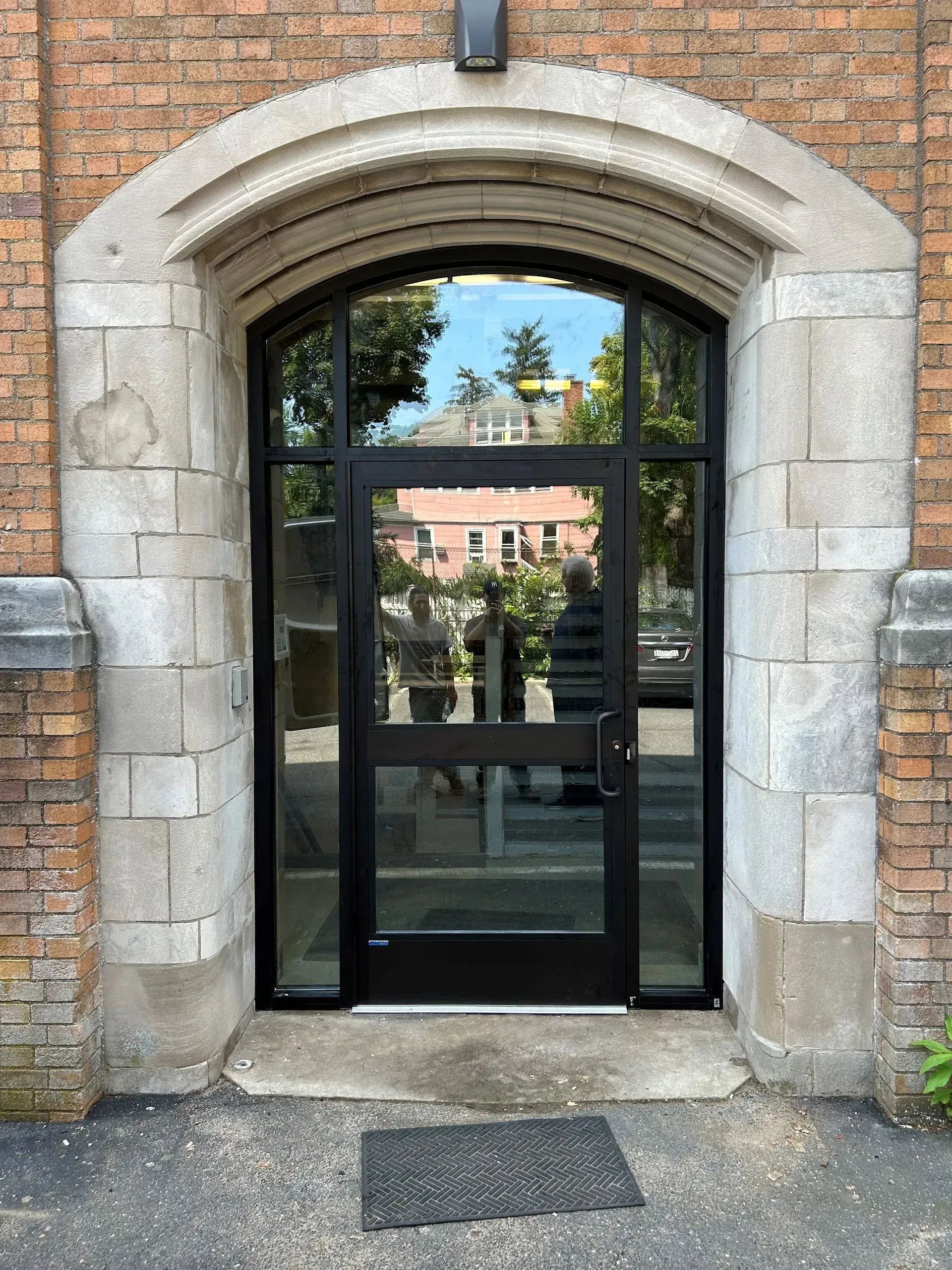 Black-framed glass door with sidelights in arched stone doorway, set in a brick building.